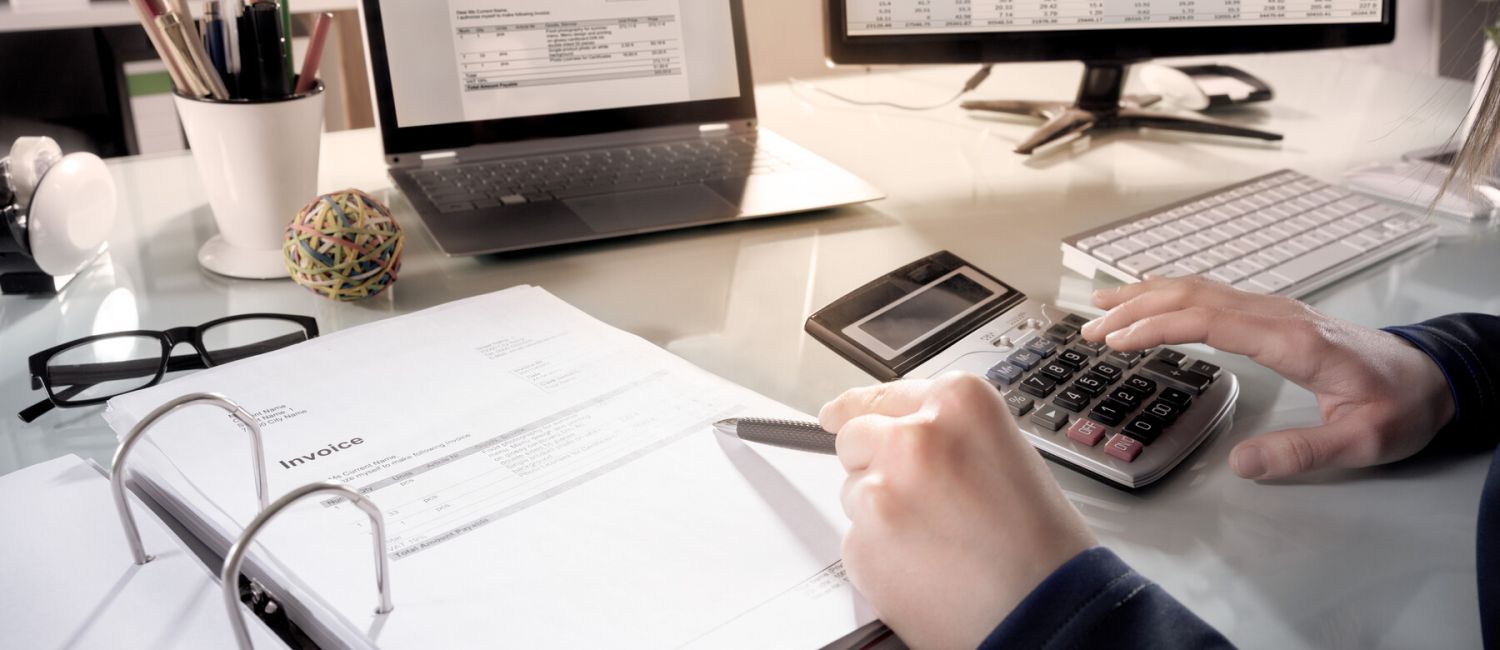 Символическое изображение: письменный стол, за которым сотрудник проверяет документы, набирает текст на калькуляторе, а перед ним стоит открытый ноутбук и монитор.